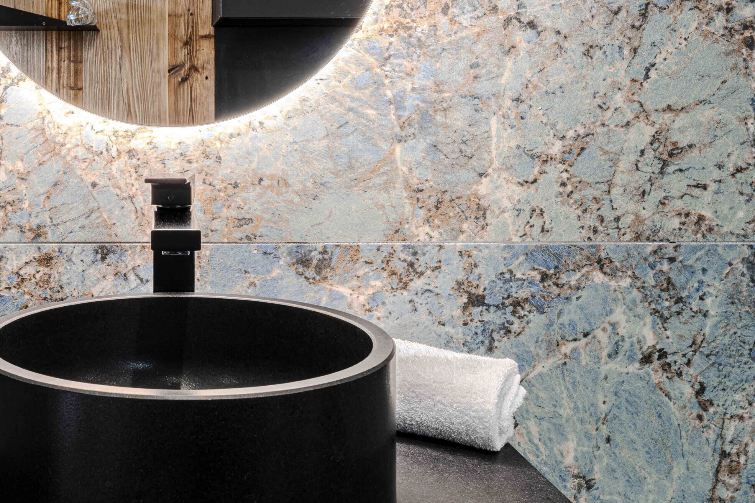 Un baño moderno con un lavabo redondo negro, un estante de madera y una pared de mármol.