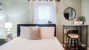 Ein ordentlich eingerichtetes Schlafzimmer mit einem Bett, einem Nachttisch mit Lampe, einem Sessel, einem Spiegel und einem Fenster mit Vorhängen.