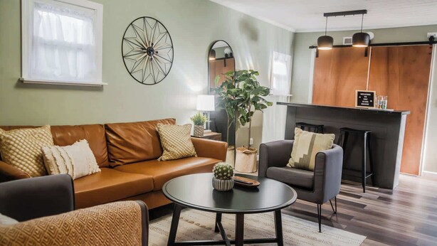 A modern living room with a brown leather sofa, a round coffee table, and a grey armchair. There is a large wall clock, a mirror, and a fireplace with a welcome sign. - Tiny Home fit for a King: Near Belmont Park! (Dayton)