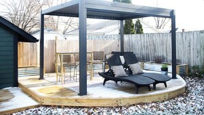 A covered patio area with a wooden deck, a table with chairs, and a lounge chair with cushions. - Tiny Home fit for a King: Near Belmont Park! (Dayton)