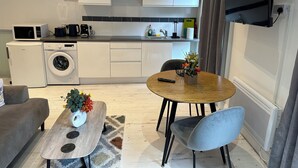 Una cocina moderna con armarios blancos, un microondas, una cafetera y una lavadora. Hay una zona de comedor con una mesa redonda, sillas y un jarrón con flores.
