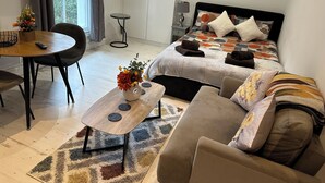 Un dormitorio moderno con cama, sofá, mesa de comedor y un jarrón de flores, junto a una mesita auxiliar.