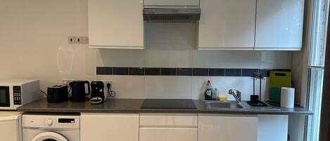 Una cocina compacta con armarios blancos, microondas integrado, cafetera y lavadora.
