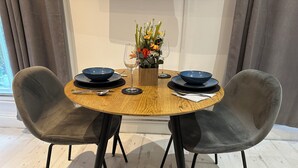 Una mesa redonda de madera con dos sillas oscuras, un jarrón de flores y una copa de vino.