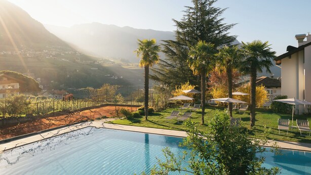 A poolside area with lounge chairs and umbrellas, surrounded by greenery and a mountainous backdrop. - Apartment with large south-facing terrace, incl. garage parking space (Dorf Tirol)