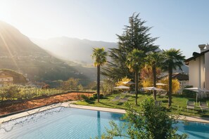 A poolside area with lounge chairs and umbrellas, surrounded by greenery and a mountainous backdrop. - Apartment with large south-facing terrace, incl. garage parking space (Dorf Tirol)