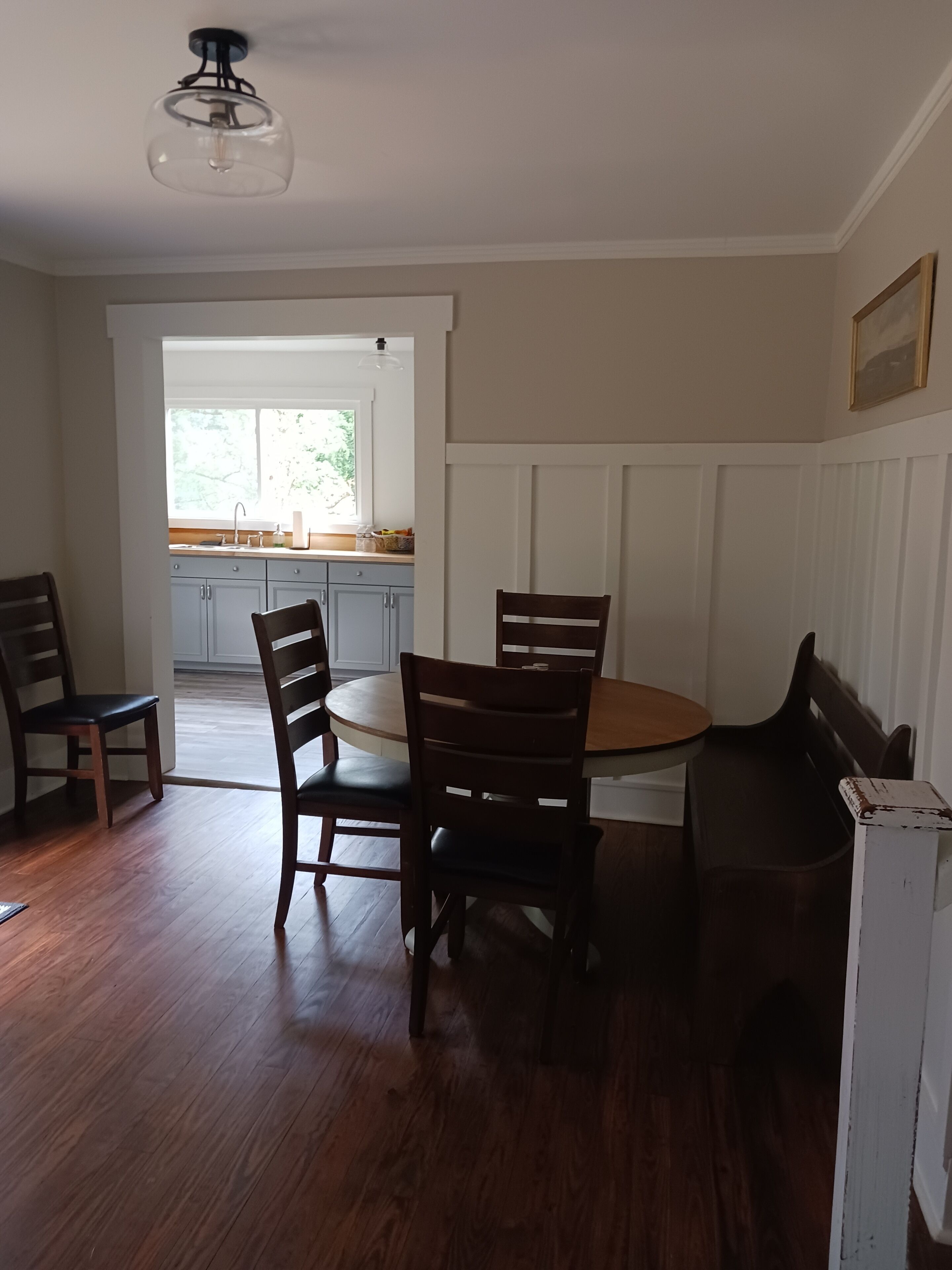 A dining area with a round table and four chairs, wooden flooring, a ceiling light fixture, and a view of a kitchen with cabinets and a window.