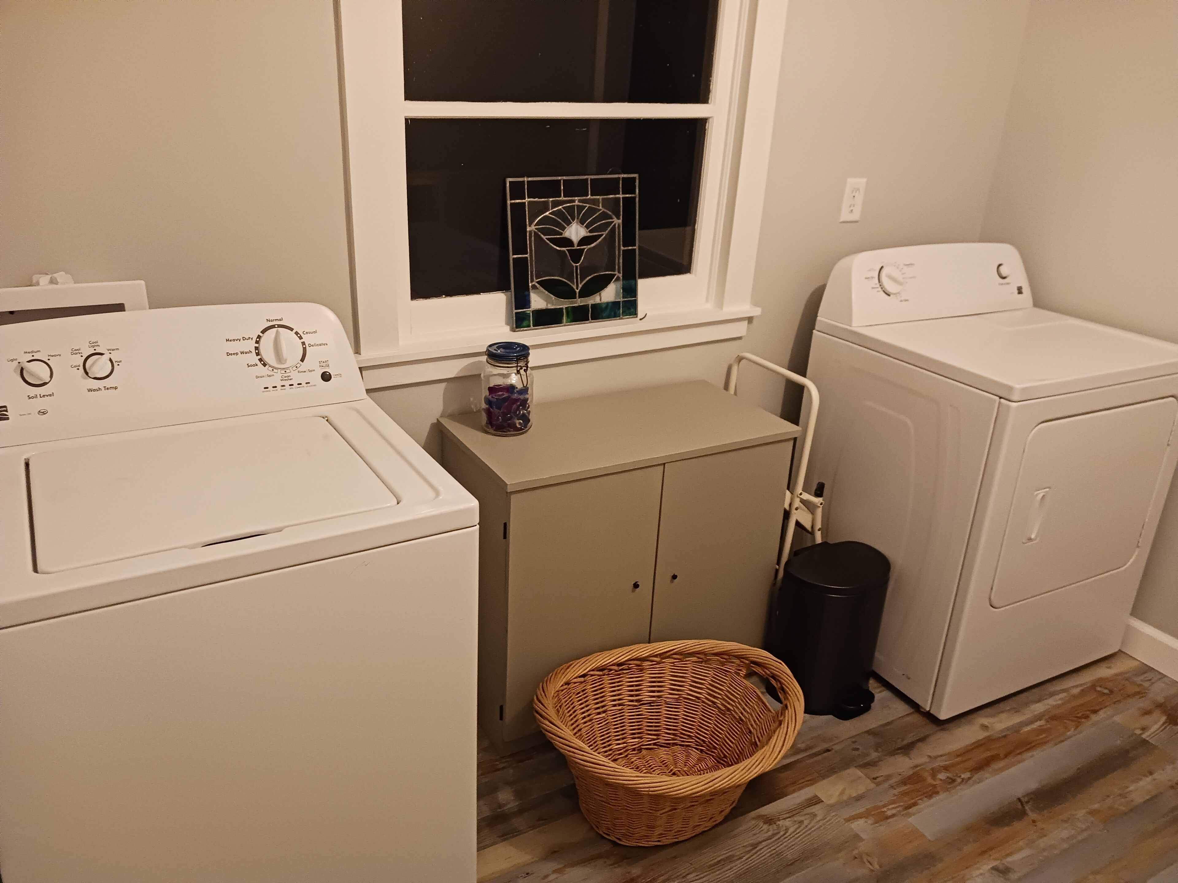 A laundry room with two washing machines, a small cabinet, a wicker basket, and a window with a decorative item.