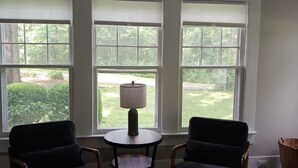 Una habitación con tres ventanas amplias, un ventilador de techo, dos sillones oscuros, una mesita redonda con una lámpara y una cesta de mimbre.