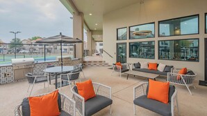 Ein möblierter Patio-Bereich im Freien mit Korbmöbeln, einem Grill und großen Fenstern mit Blick auf einen Tennisplatz.