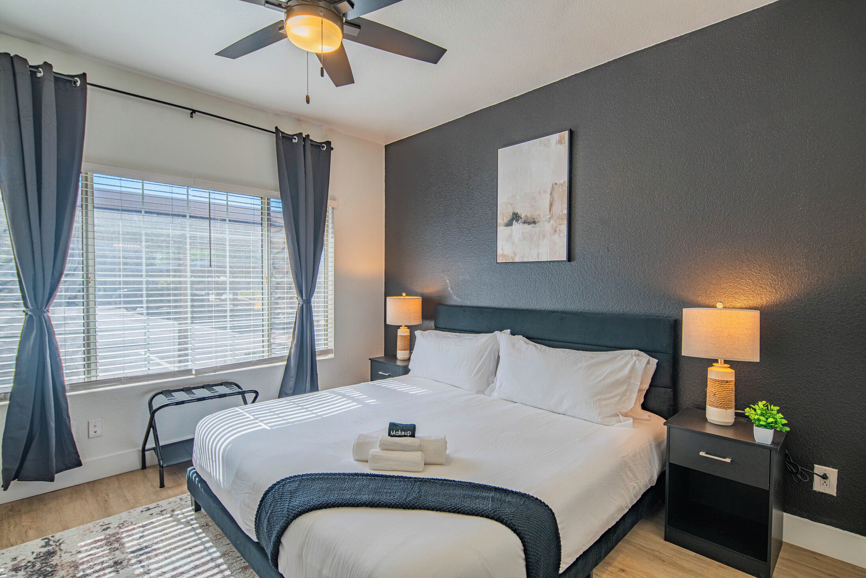 A modern hotel room with a large bed, dark headboard, bedside lamps, a small table with a lamp, and a view of the outside through a window with curtains.