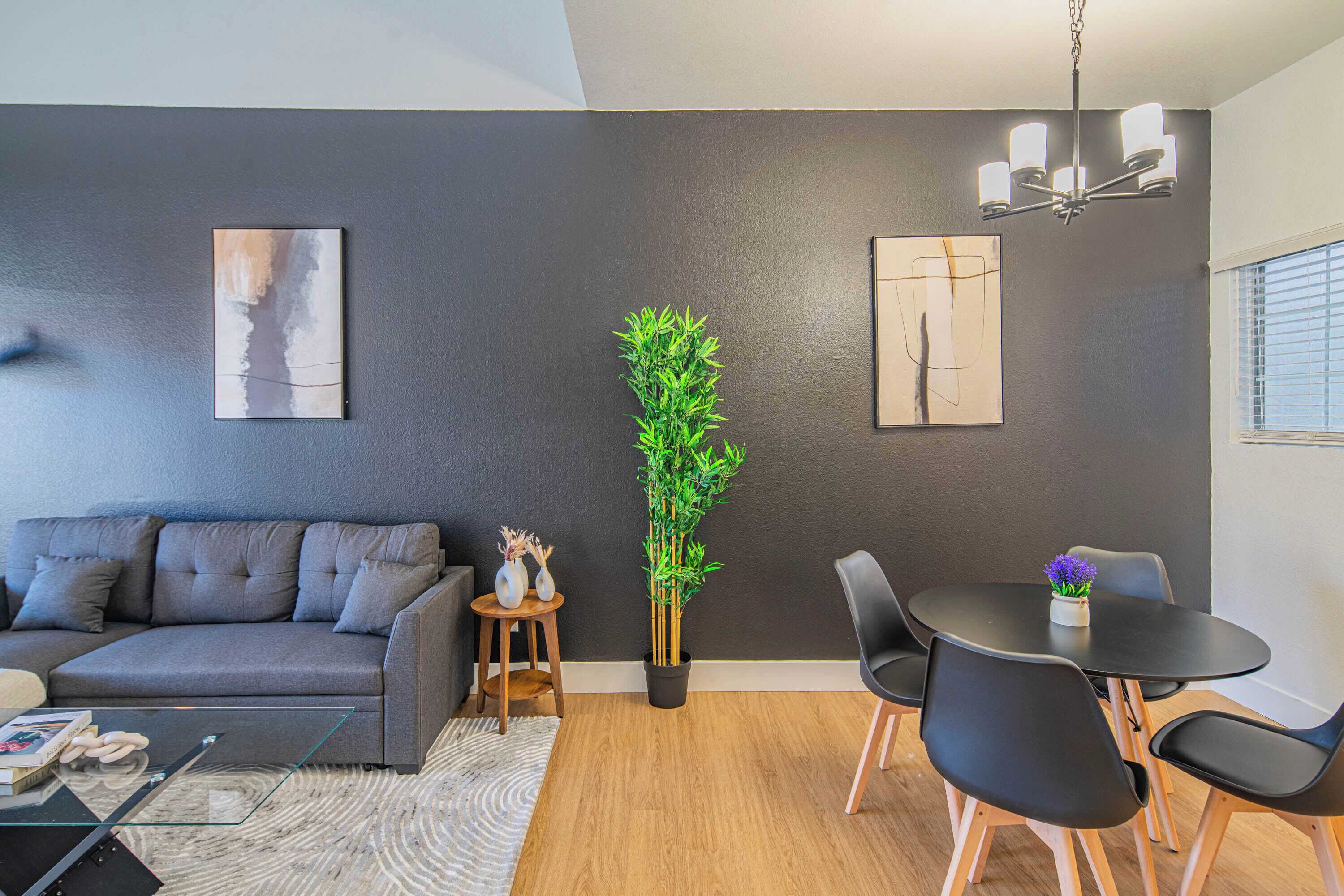 A modern living room with a grey sofa, a dining table with chairs, a potted plant, and artwork on the walls.