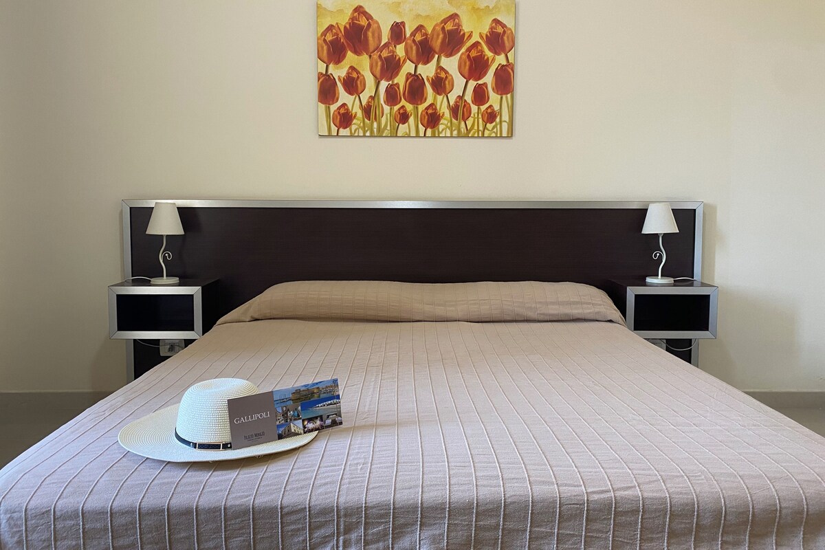 A hotel room with a large bed, two bedside tables with lamps, a wall-mounted painting of flowers, a hat, and a brochure on the bed.