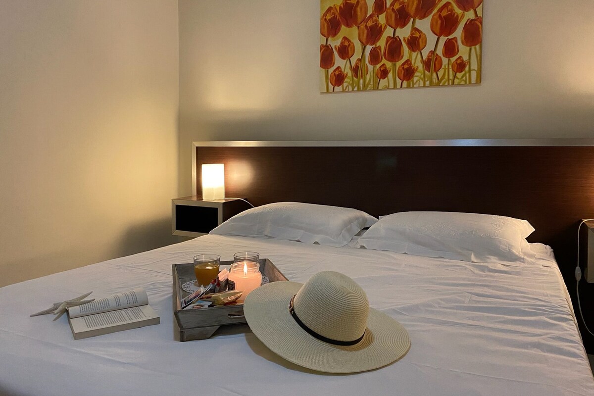 Una camera d'albergo con un letto rifatto, un comodino con una lampada, un vassoio con un libro, una candela e un cappello.