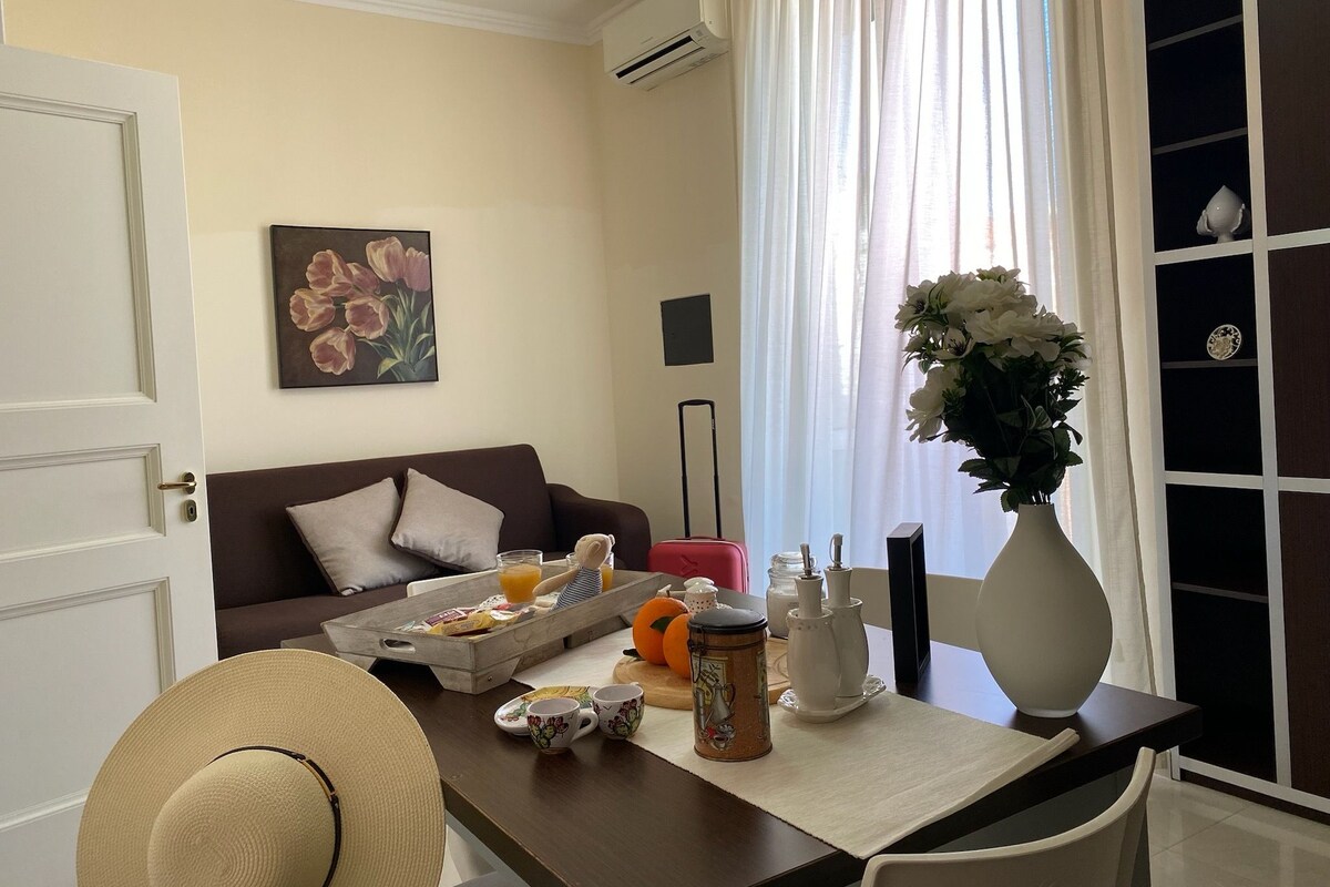 A hotel room with a dining table set with breakfast items, a sofa, a painting, and a vase with flowers.