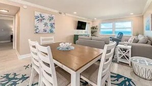 Uma sala ampla com uma mesa de jantar de madeira, cadeiras brancas, um sofá cinza e uma TV sobre um móvel.
