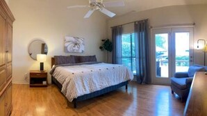 Ein Schlafzimmer mit Bett, Nachttisch, Deckenventilator, Fenster mit Vorhängen und Blick ins Freie.