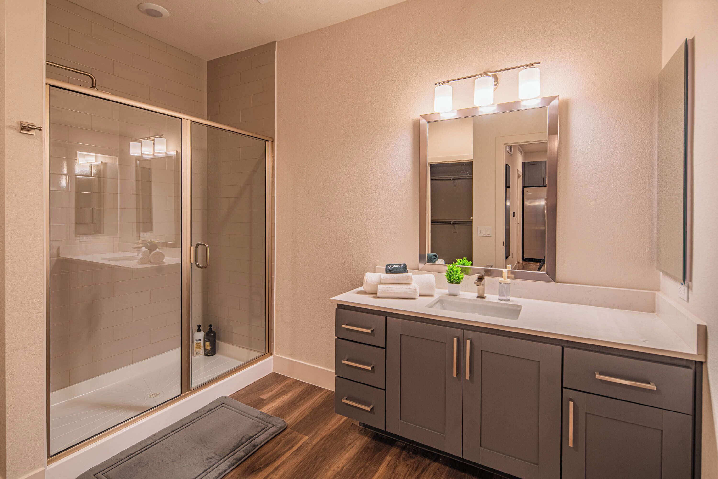 Une salle de bain moderne dotée d’une douche à parois de verre, d’un lavabo avec un miroir au-dessus et d’un meuble-lavabo aux tons foncés, muni de tiroirs.