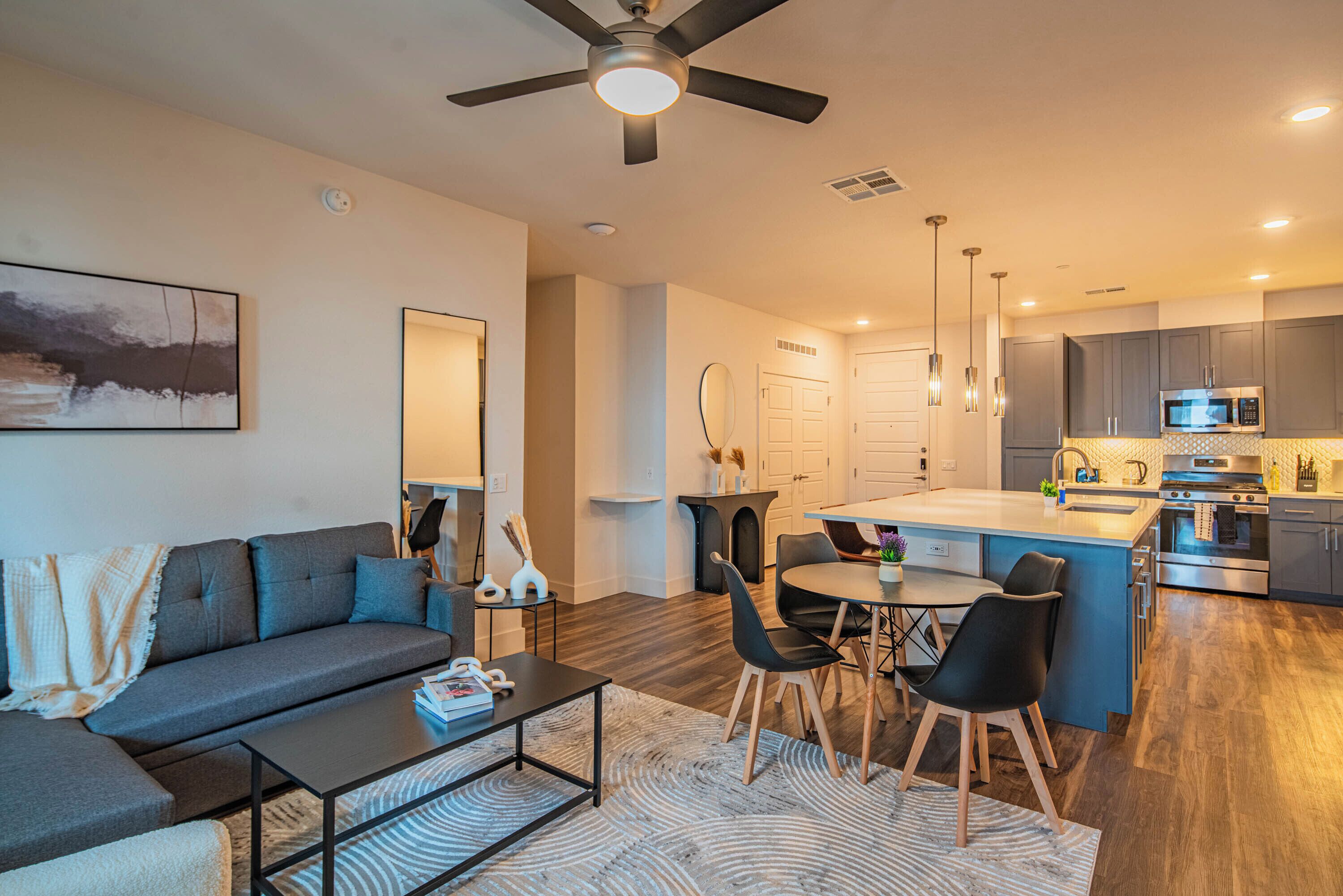 A modern living room with a sofa, coffee table, and dining area. The kitchen features a central island, microwave, and stainless steel appliances.