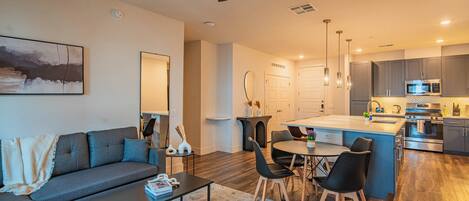 A modern living room with a sofa, coffee table, and dining area. The kitchen features a central island, microwave, and stainless steel appliances.