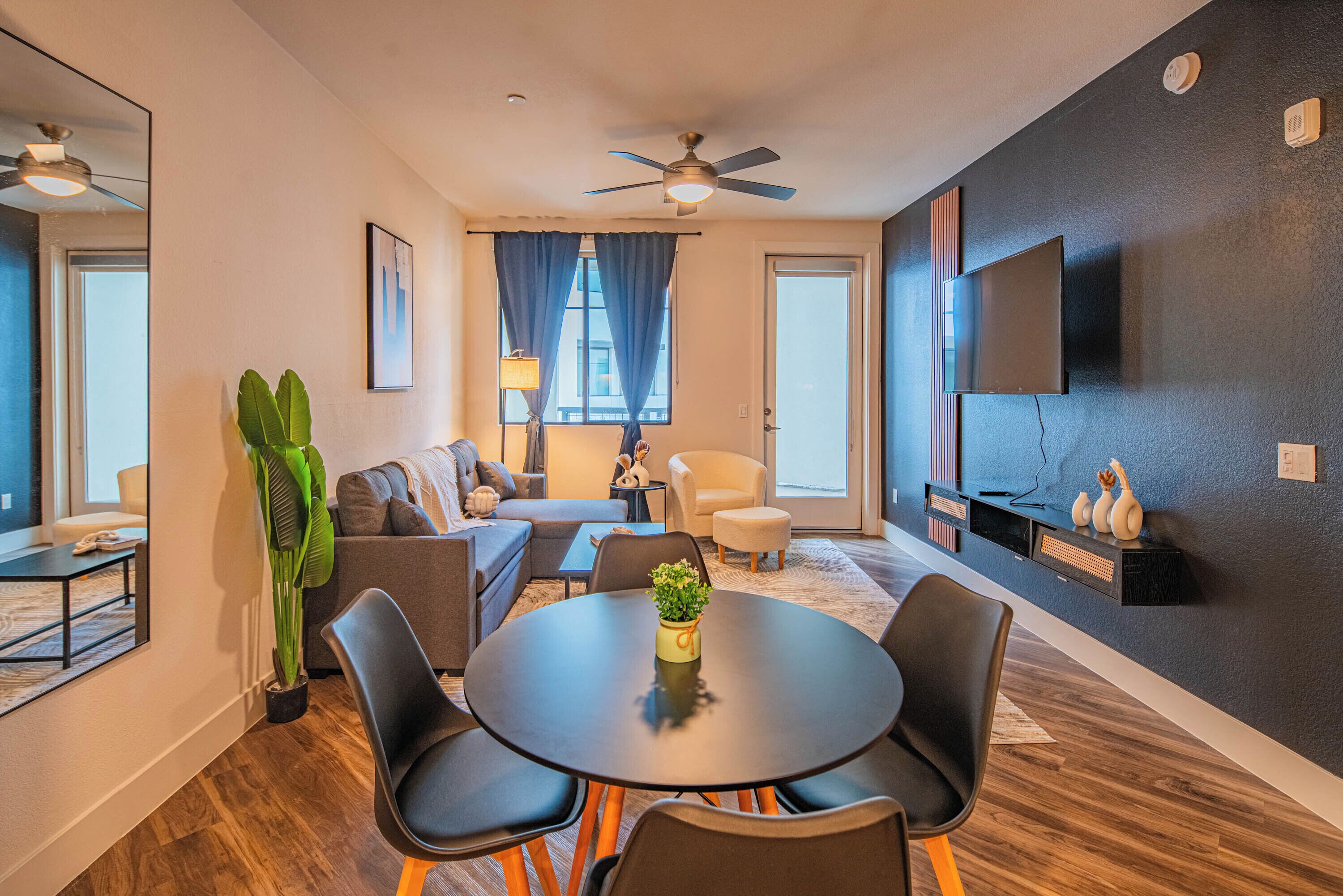 A modern living room with a round table, black chairs, a sofa, a television, and a ceiling fan.