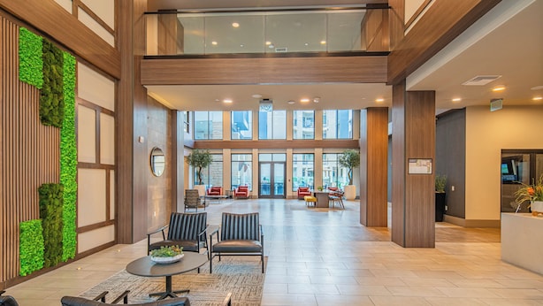 Eine großzügige Lobby mit hoher Decke, Holzvertäfelung und einem vertikalen Grünbereich. Mehrere Sitzbereiche mit Stühlen und Tischen laden zum Verweilen ein, im Hintergrund ist eine Rezeption erkennbar.