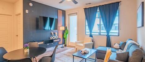 A modern living room with a flat-screen TV, a ceiling fan, a dining area with a table and chairs, and a sofa with a throw blanket.
