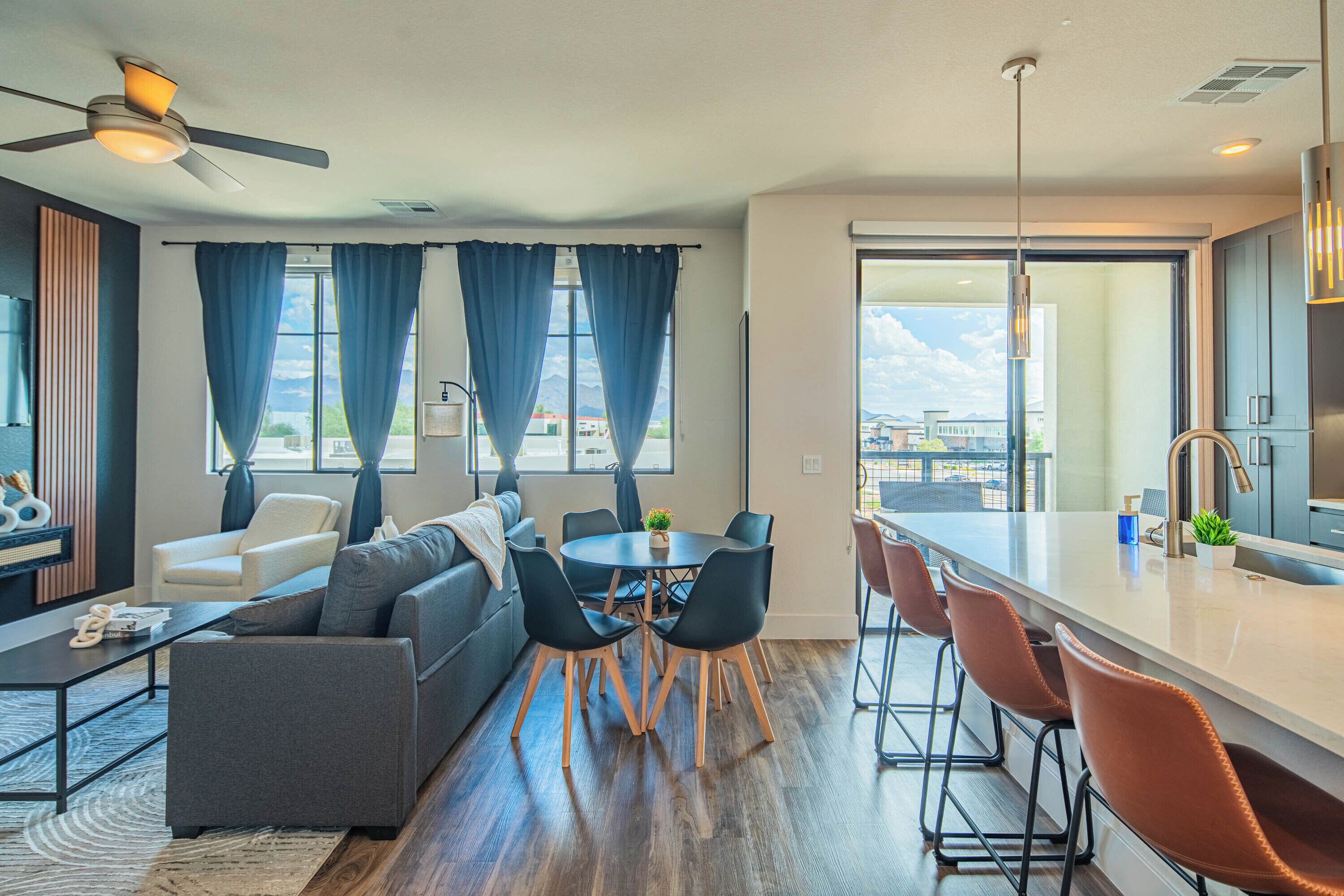 A modern living room with a sofa, dining table, and chairs, a kitchen area with a bar, and large windows with curtains.