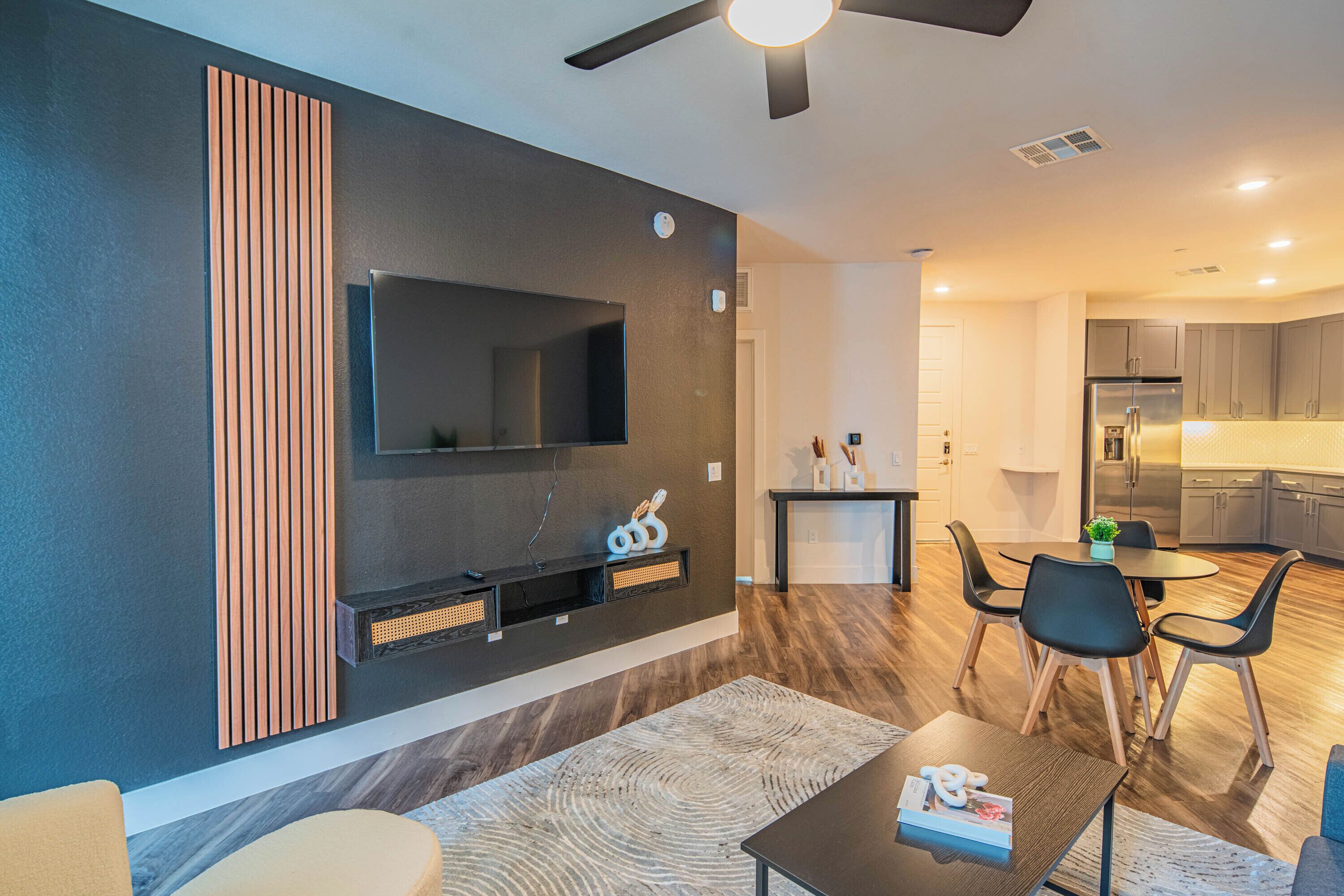 A modern living room with a flat-screen TV, a dining area with a table and chairs, and a kitchen in the background.