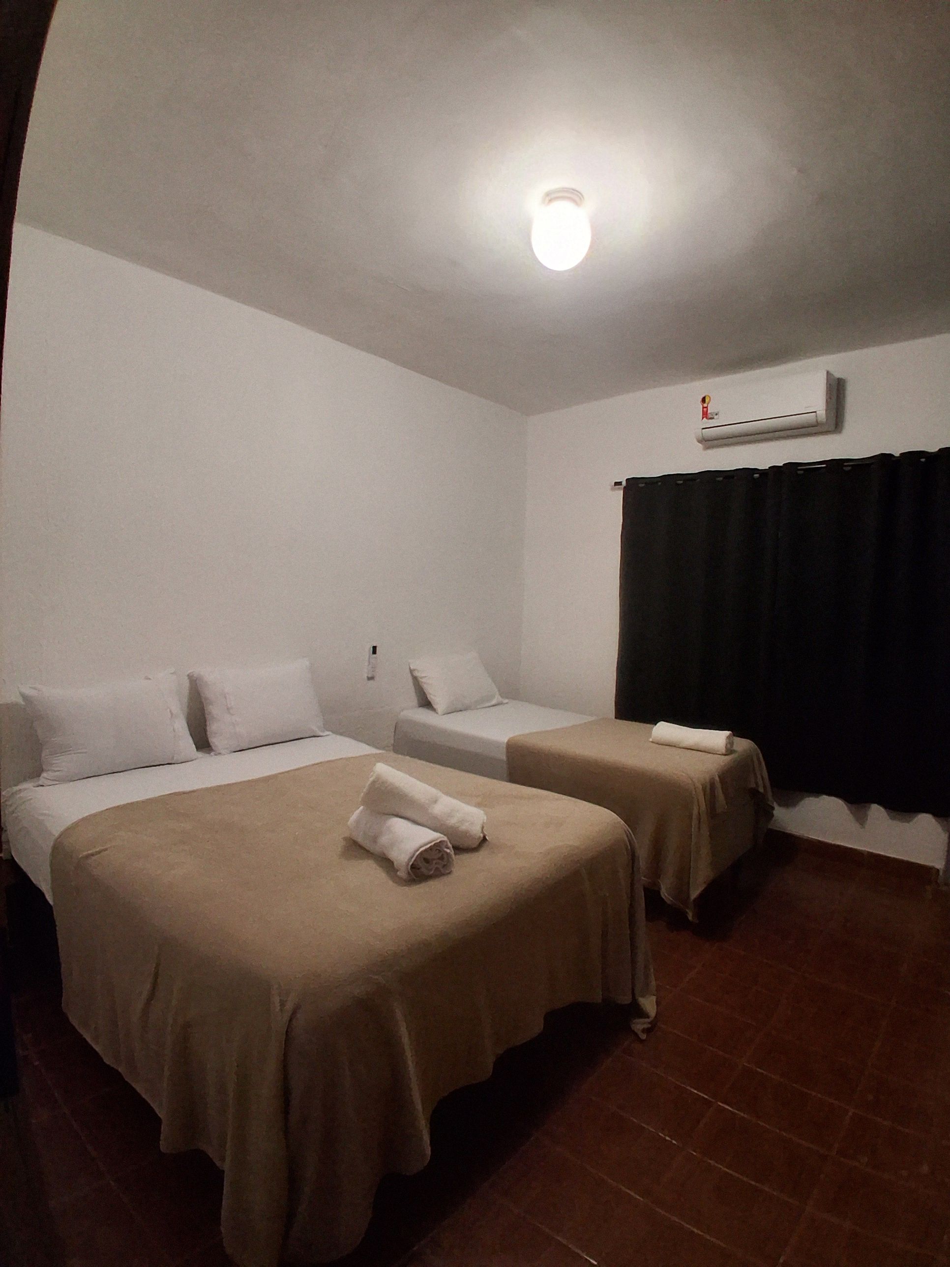 A hotel room with two beds, a ceiling light, an air conditioning unit, and a black curtain.