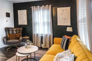 Un salon avec un fauteuil en cuir, un canapé jaune, une table basse ronde et des œuvres d’art abstraites accrochées aux murs.