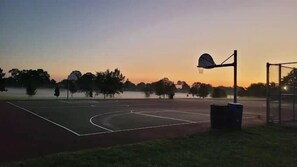 Een buitenbasketbalveld in de schemering, met een basketbalring, een prullenbak en een hek op de achtergrond.