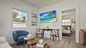 Una sala de estar moderna con un cuadro grande de temática playera, un sillón azul, una mesa de centro de madera y vista al dormitorio por una puerta abierta.