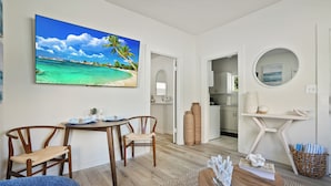 Una sala de estar con un cuadro grande de playa, muebles de madera, un espejo redondo y una cocina al fondo.