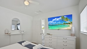 Un dormitorio con cama, televisor en la pared, cómoda y puerta que da a otra habitación.