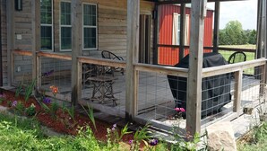 Een houten blokhut met een overdekte veranda, een barbecue en een tuin vol bloemen en planten.