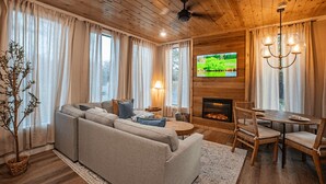 A cozy living room with a sofa, a fireplace, a dining table with chairs, and a television mounted on the wall.