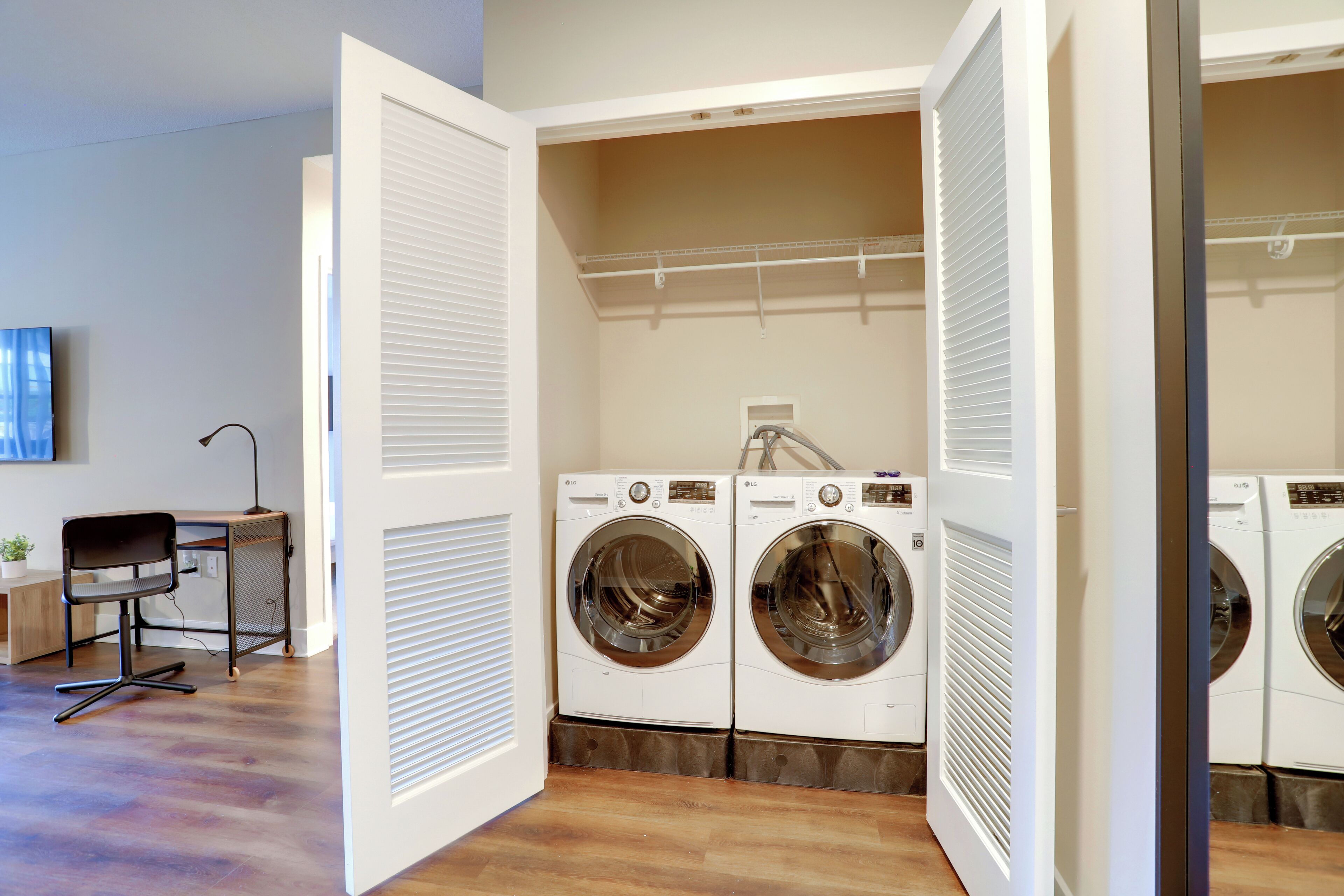 A laundry room with two front-loading washing machines, a desk with a chair, and a television on the wall.