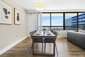 Un espace de repas moderne avec une table en bois dressée pour quatre personnes, entourée de chaises en osier. Une grande fenêtre offre une vue sur un immeuble et un balcon.