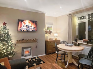 Un salon chaleureux avec une cheminée, un téléviseur diffusant une image de feu de cheminée, un sapin de Noël et un coin repas avec des chaises et une table.