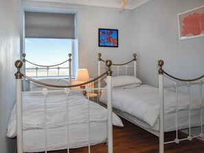 A room with two white metal beds, a window with a view, a lamp on a bedside table, and a framed picture on the wall. - Marina Views, your peaceful coastal escape in the heart of Amble. (Morpeth)