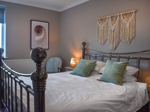 A bedroom with a bed, a chair, a radiator, a wall-mounted macrame piece, and a framed picture.