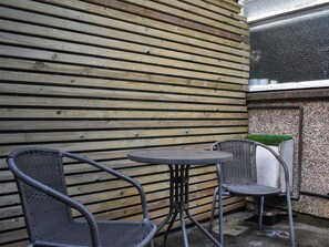Pared de madera con tablones horizontales, una mesa redonda de metal y dos sillas de mimbre.