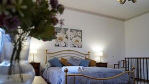 Un dormitorio con cama, dos mesitas de noche, una mesita auxiliar, un jarrón con flores y dos cuadros de flores colgados en la pared.