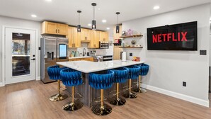 Une cuisine moderne dotée d’un îlot central, de tabourets de bar et d’un téléviseur fixé au mur qui diffuse du contenu « NETFLIX ».