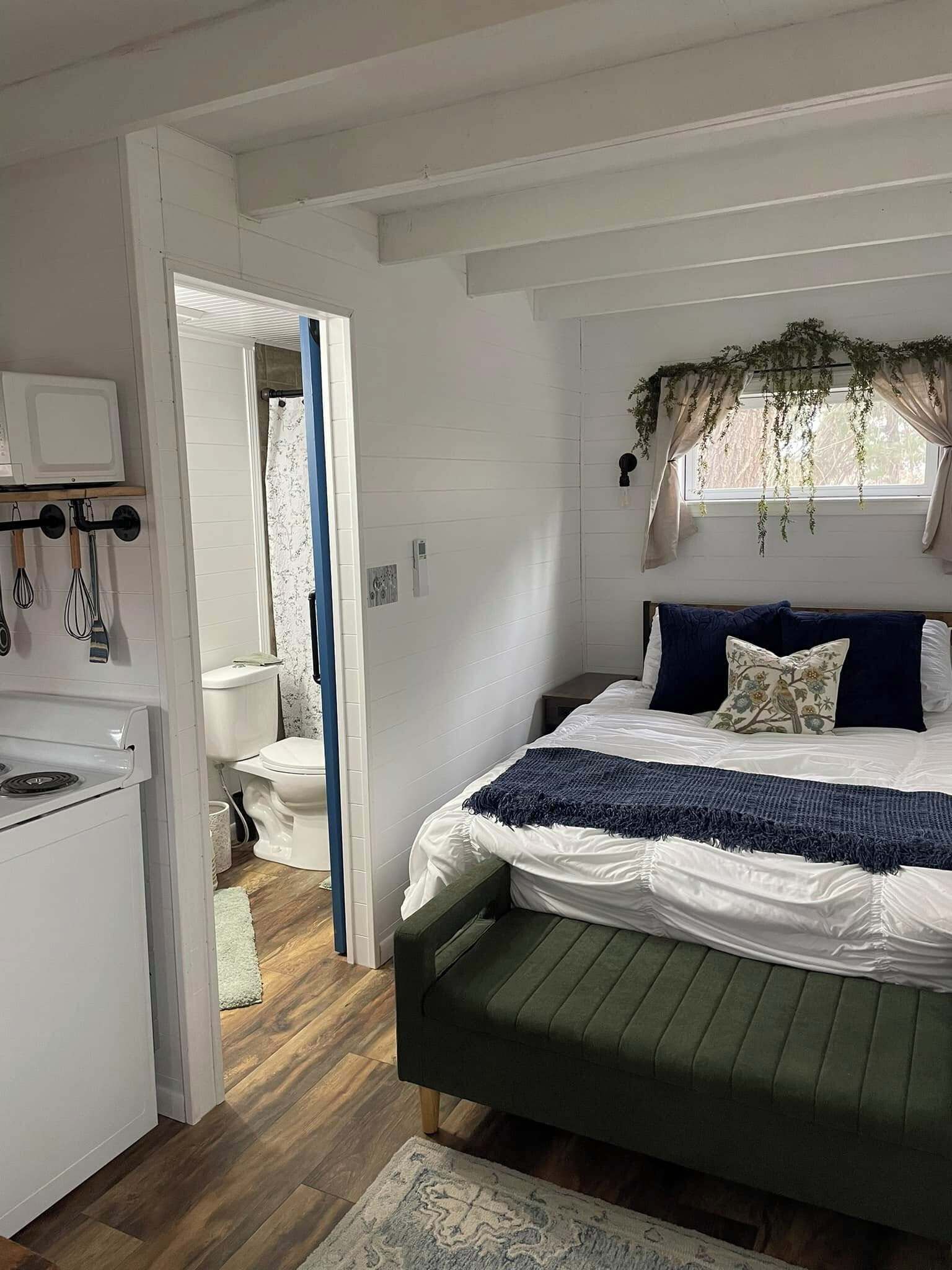 A bedroom with a bed, a window with curtains, a hanging plant, and a kitchenette area.