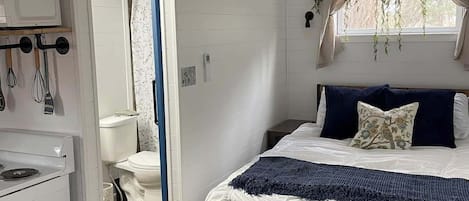 A bedroom with a bed, a window with curtains, a hanging plant, and a kitchenette area.