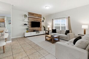 Ein modernes Wohnzimmer mit Flachbildfernseher, weißen Sofas, einem Couchtisch und einem Essbereich mit Stühlen und Tisch.