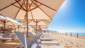 A beachside hotel with rows of sun loungers under large umbrellas, a sandy beach, and the ocean in the background.