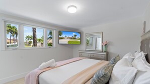 A bedroom with a bed, a TV mounted on the wall, a dresser, and a window with a view of palm trees and a house.